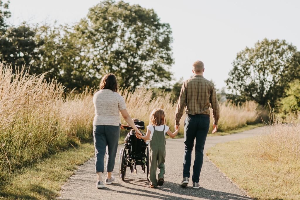 A mother and father take a walk with their 2 kids, one of whom uses a wheelchair, outdoors on a paved path through a prairie
