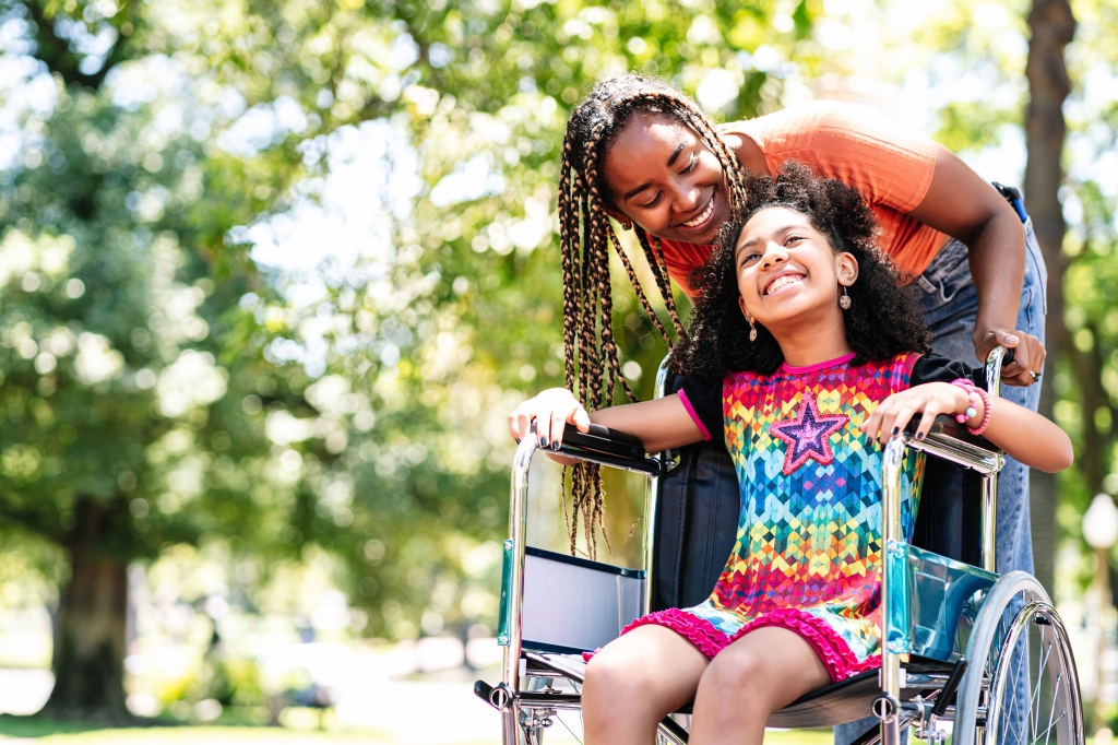 A smiling young mother or caregiver pushes a happy girl in a wheelchair, outdoors in nature
