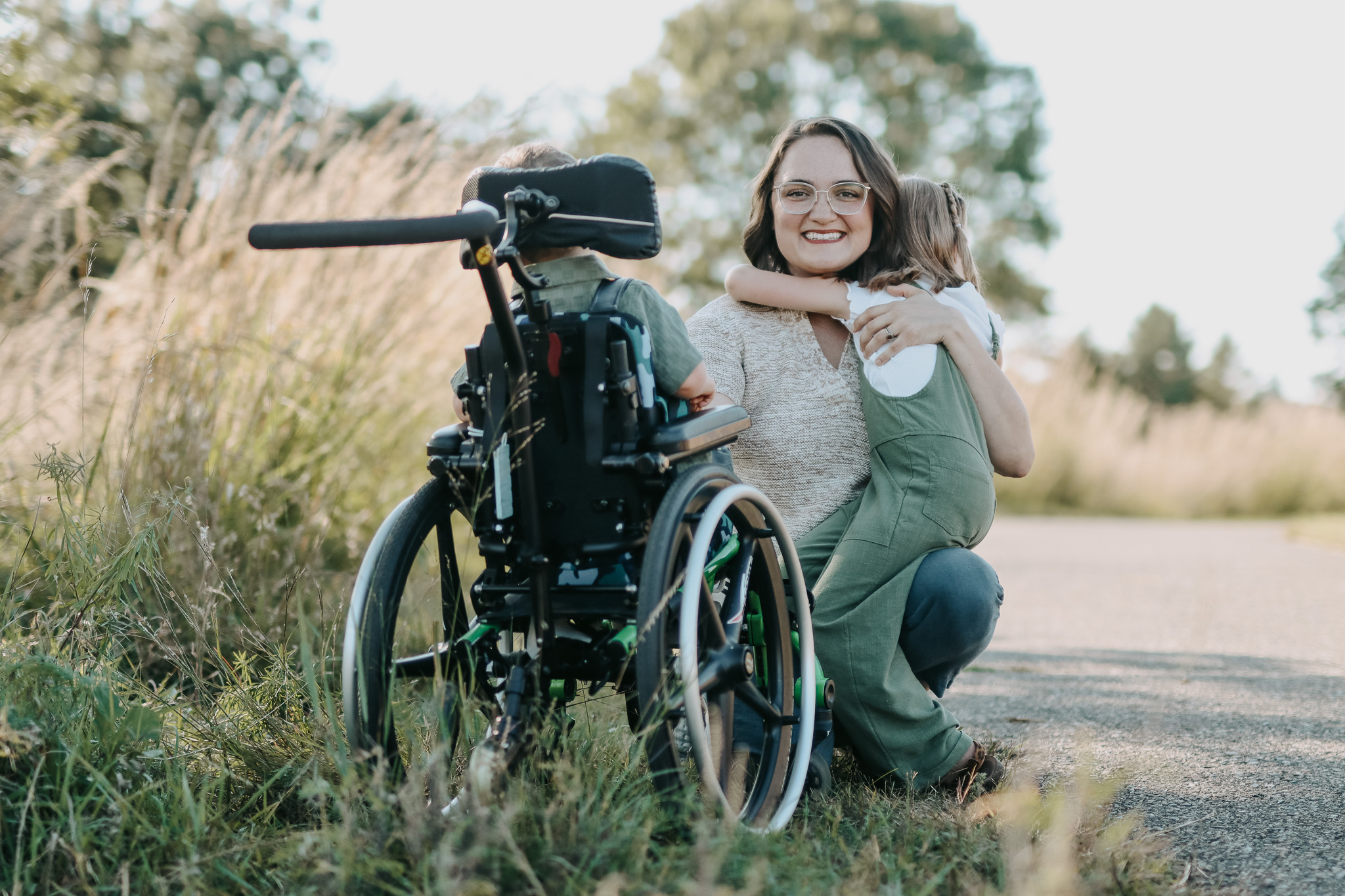 A mother smiles for the camera while embracing her two young children, one of whom is in a wheelchair, outdoors in nature